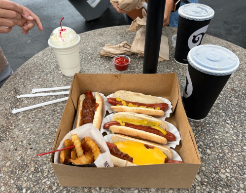 A box of hot dogs with toppings, fries on a stick, and drinks on a table with condiments.