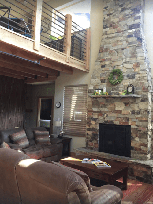 Cozy living room with a stone fireplace, wooden beams, and a loft area, featuring comfortable seating and natural light.