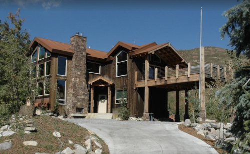 A modern wooden house with large windows, a stone chimney, and a deck, surrounded by trees and rocky landscaping.