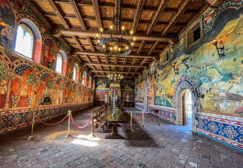 A grand dining hall with ornate wooden beams, colorful murals, and a long table, illuminated by chandeliers.