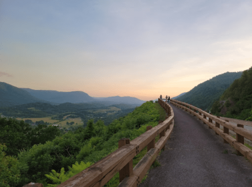A scenic pathway with a wooden railing, overlooking a valley and mountains at sunset, with two people walking ahead.