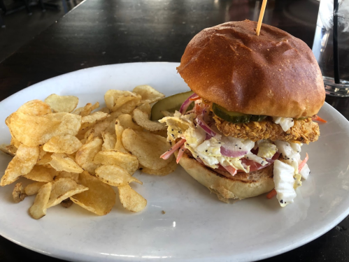 A crispy chicken sandwich with coleslaw and pickles, served with a side of potato chips on a white plate.