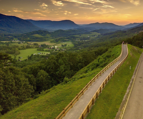 A winding path along a hillside overlooks a lush green valley at sunset, with mountains in the background.