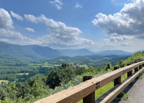 A scenic view of rolling green hills and mountains under a blue sky with fluffy clouds, framed by a wooden railing.