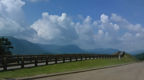 A scenic view of rolling hills and mountains under a blue sky with fluffy clouds, alongside a winding road and wooden fence.