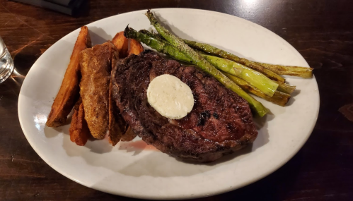 A plate featuring a grilled steak topped with butter, served with sweet potato fries and grilled asparagus.