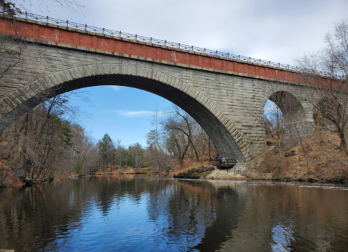 A stone arch bridge spans over a calm river, surrounded by trees and a clear blue sky.
