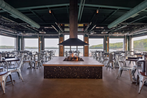 Modern restaurant interior with a central fireplace, surrounded by tables and large windows overlooking a scenic view.
