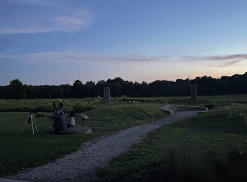 A serene landscape at dusk with a path, telescope, and two figures observing the sky in a grassy area.