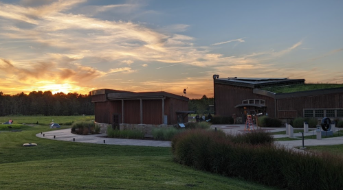 A sunset over a modern building surrounded by green grass and trees, with a clear sky and scattered clouds.