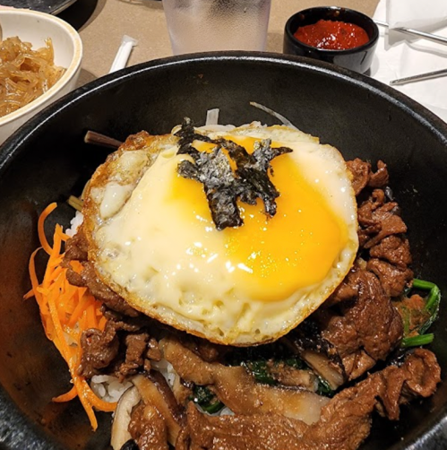 A sizzling stone bowl filled with beef, vegetables, and a fried egg topped with seaweed, served with sauce on the side.