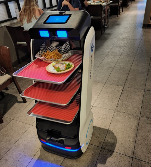 A food delivery robot with three trays, carrying a plate of fish and fries, moves through a restaurant.