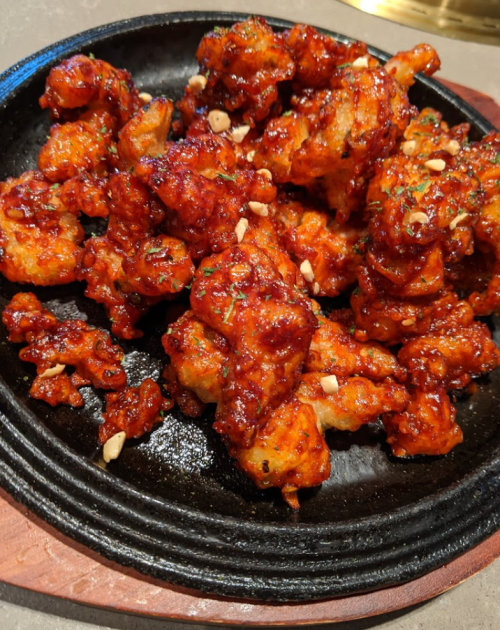 A sizzling plate of crispy, glazed chicken pieces topped with chopped peanuts and herbs.