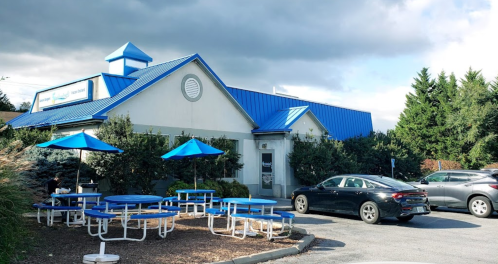 A restaurant with a blue roof, outdoor seating, and parked cars under a cloudy sky.