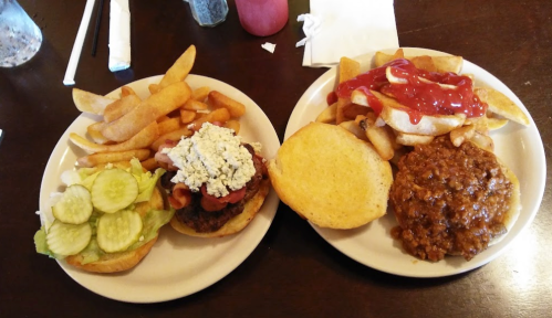 Two plates of food: one with a burger topped with blue cheese and bacon, and the other with a chili burger and fries.