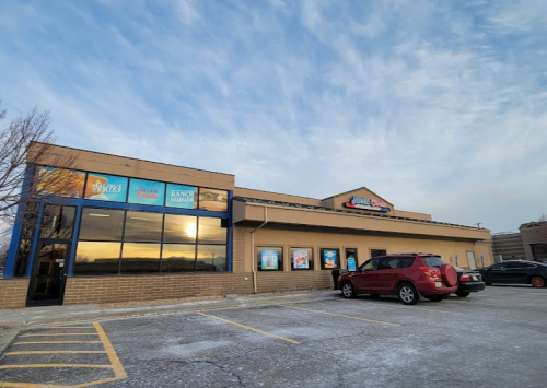 Exterior of a restaurant building with large windows and signage, parked cars in the foreground, under a cloudy sky.