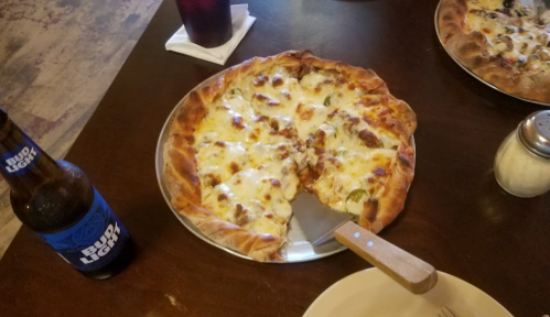 A pizza with melted cheese and toppings on a metal tray, alongside a bottle of Bud Light and a salt shaker.