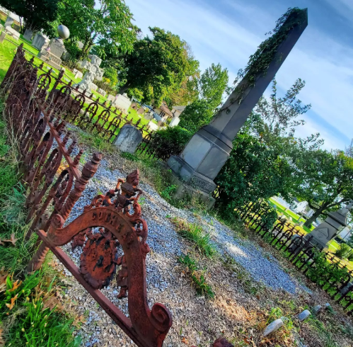 A weathered cemetery with a tall gravestone, surrounded by an ornate iron fence and overgrown greenery.