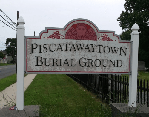 Sign for Piscatawaytown Burial Ground, featuring an angel design, with a grassy area and road in the background.