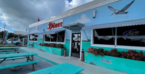 A colorful diner with a mural, outdoor seating, and flower beds, set against a cloudy sky.