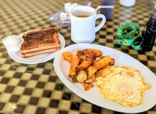 A plate with fried eggs, seasoned potatoes, and toast, alongside a cup of coffee on a checkered table.