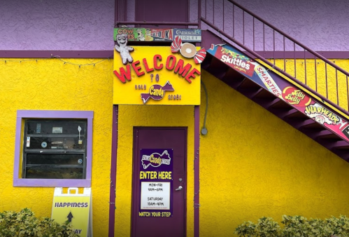 Colorful candy store entrance with a bright yellow exterior, purple accents, and a welcoming sign. Stairs lead to the upper level.