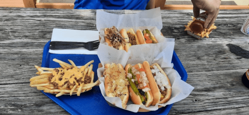 A tray with four hot dogs topped with various ingredients, a side of loaded fries, and a hand reaching for a condiment.