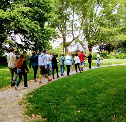 A group of people walking along a path in a lush green park, surrounded by trees and a house in the background.