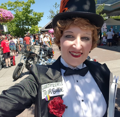 A person in a tuxedo and top hat smiles for a selfie, holding a clipboard at a lively outdoor event.