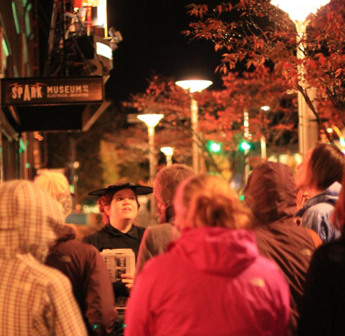 A group of people gathers at night, listening to a speaker wearing a hat outside a museum, surrounded by autumn leaves.