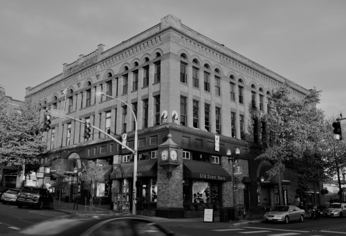 Historic brick building on a street corner, featuring large windows and a clock, captured in black and white.