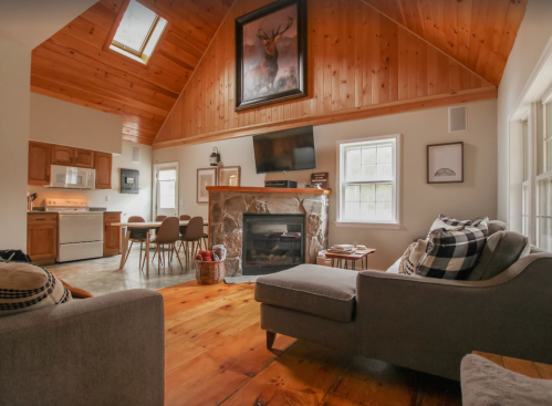 Cozy living room with wooden accents, a stone fireplace, and a dining area, featuring large windows and natural light.