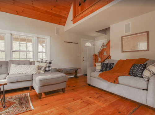 Cozy living room with gray sofas, wooden floors, and a warm throw blanket, featuring natural light and rustic decor.