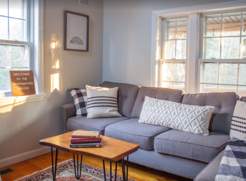 Cozy living room with a gray sofa, decorative pillows, a wooden coffee table, and natural light from large windows.