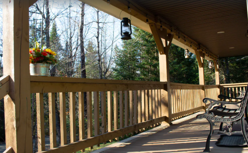A wooden porch with benches, lanterns, and a flower arrangement, surrounded by trees in a serene outdoor setting.