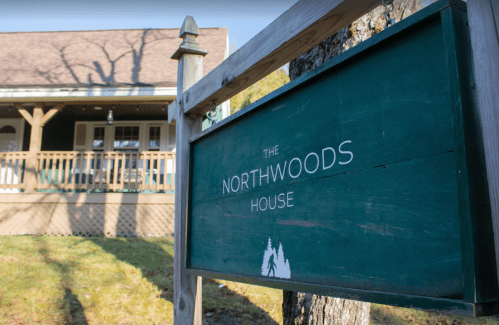 Sign for "The Northwoods House" in front of a cozy cabin with a porch and trees in the background.