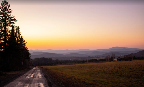 A winding dirt road leads through a serene landscape at sunset, with rolling hills and a clear sky in the background.