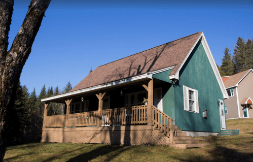 A green house with a porch, surrounded by trees and a clear blue sky, set in a rural landscape.