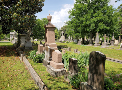A peaceful cemetery scene with various gravestones surrounded by greenery and trees under a partly cloudy sky.