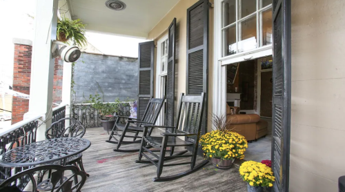 A cozy balcony with rocking chairs, a small table, and potted flowers, featuring open shutters and a view of a living space.