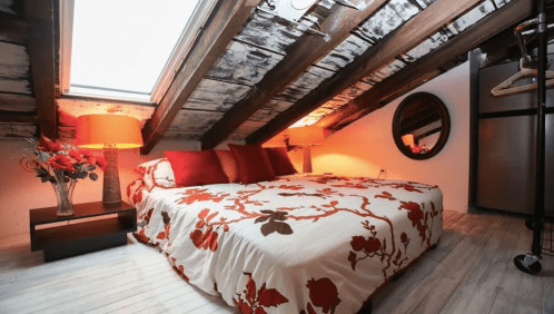 Cozy attic bedroom with a floral bedspread, warm lighting, and wooden beams, featuring a round mirror and a small table.