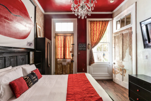 Cozy bedroom with red accents, a chandelier, and large artwork featuring lips on the wall. Bright curtains frame the window.