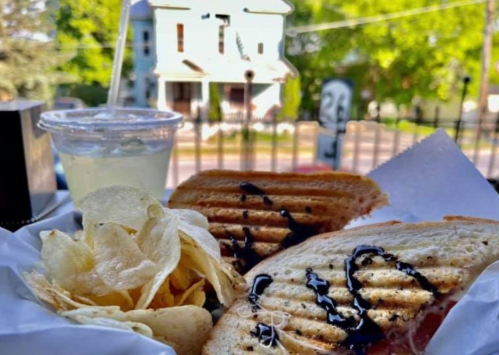 A plate with two grilled sandwiches drizzled with sauce, served with chips and a drink, with a house in the background.