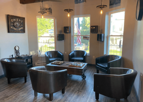 Cozy seating area with four chairs, a chess table, and large windows featuring American flags and vintage decor.