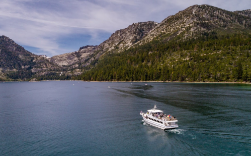 A white boat glides across a calm lake, surrounded by mountains and lush greenery under a clear sky.