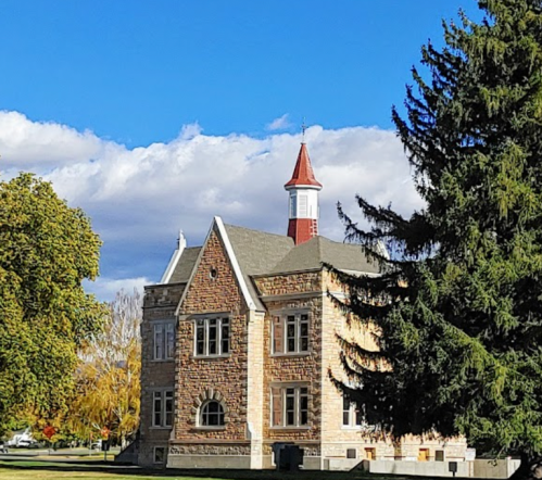 Historic stone building with a red-topped tower, surrounded by trees and a clear blue sky.