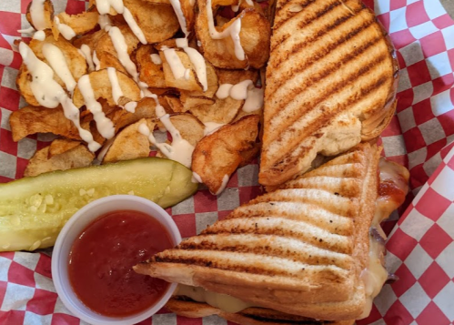A grilled sandwich with melted cheese, a pickle, potato chips drizzled with sauce, and a side of ketchup in a basket.
