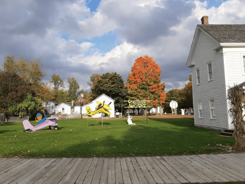 Colorful autumn scene featuring a whimsical airport sign, a cartoon plane, and a white house on a grassy lawn.