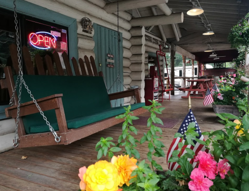 A cozy porch with a green swing, flowers, and American flags, featuring an "Open" neon sign in the background.