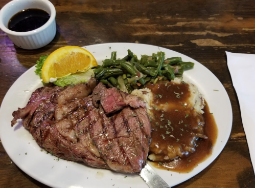 A plate with grilled steak, green beans, mashed potatoes with gravy, and a slice of orange, alongside a small bowl of sauce.